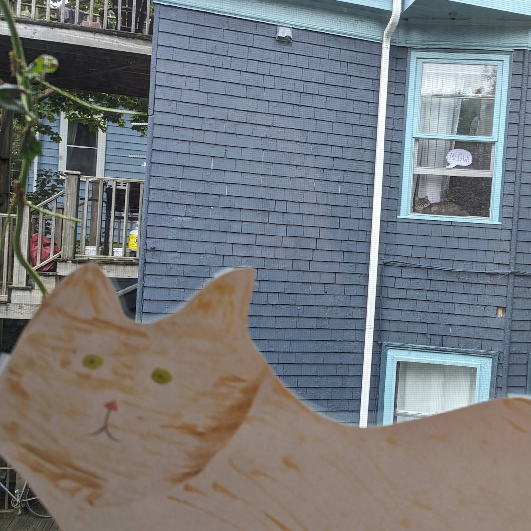 A paper cat in a window. In the background, a neighbouring cat has a speech bubble that says meow.